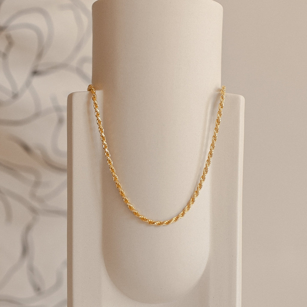 Gold rope chain necklace hanging in a graceful arc on a minimal cream ceramic vase.
