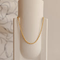 Gold rope chain necklace hanging in a graceful arc on a minimal cream ceramic vase.