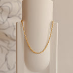 Gold rope chain necklace hanging in a graceful arc on a minimal cream ceramic vase.