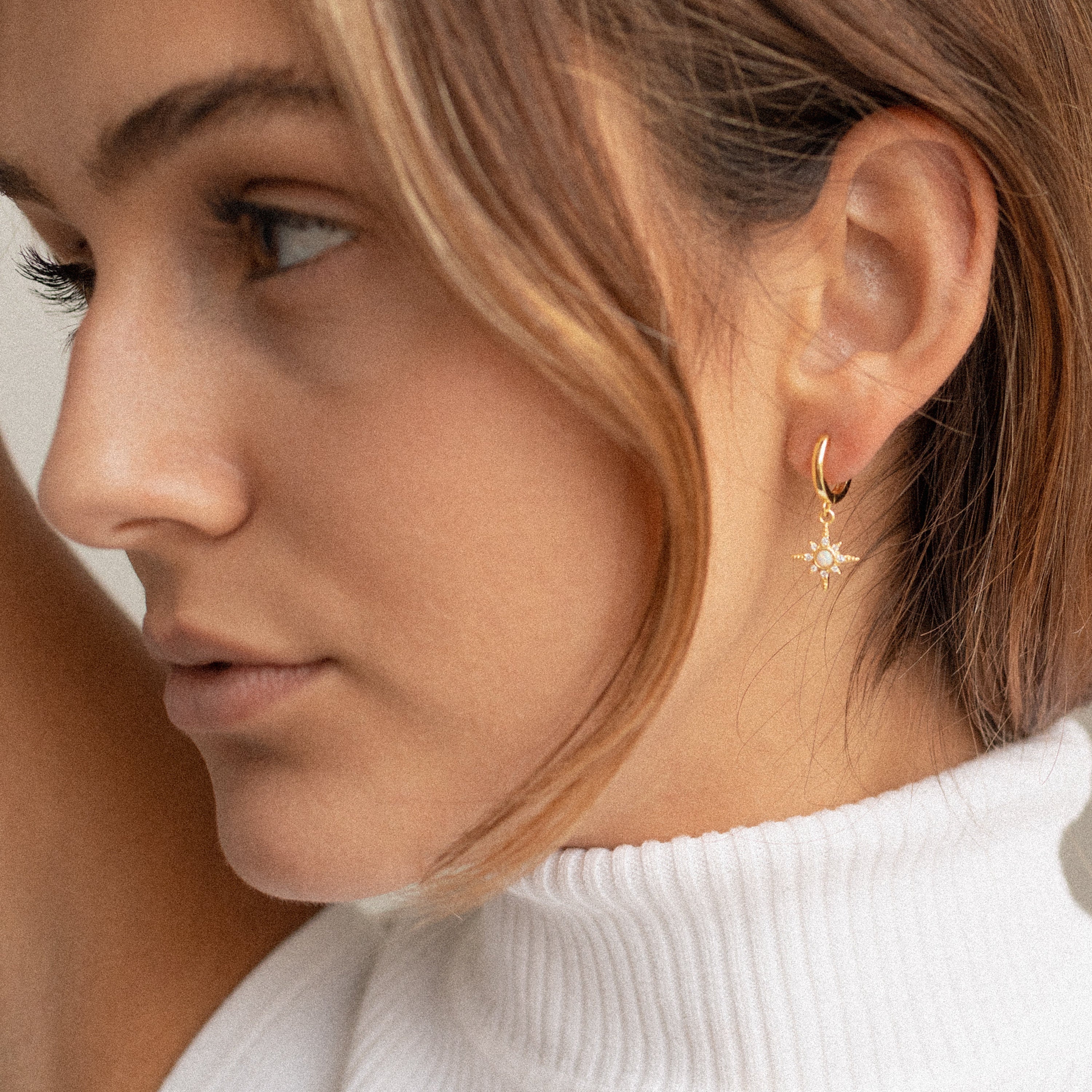 Woman with light brown hair wears a white turtleneck and elegant Starburst Huggies in Sterling Silver, accented by a gold sun-shaped earring.