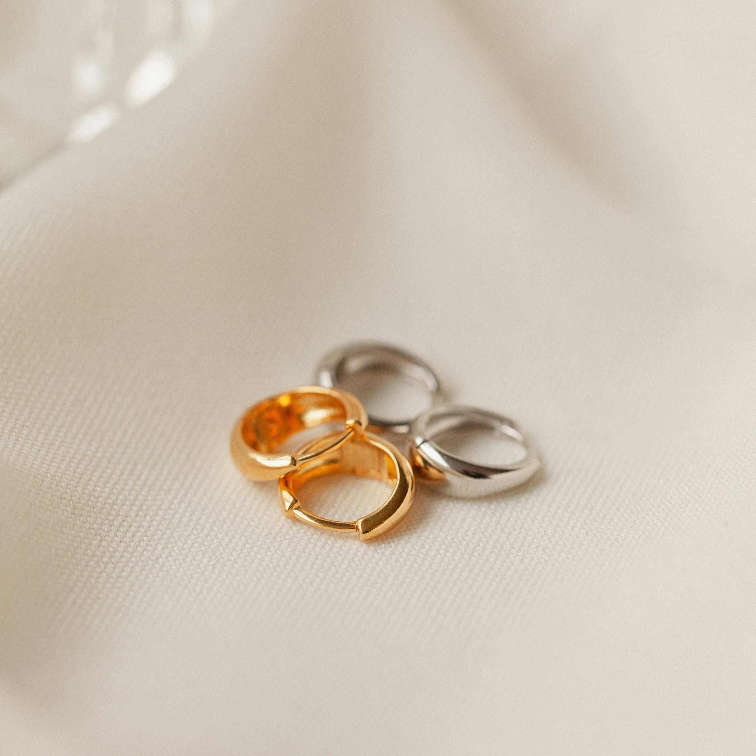 Two pairs of Lucy Hinged Huggies in Sterling Silver are displayed on a soft, cream fabric background, highlighting their minimalist hoop elegance.