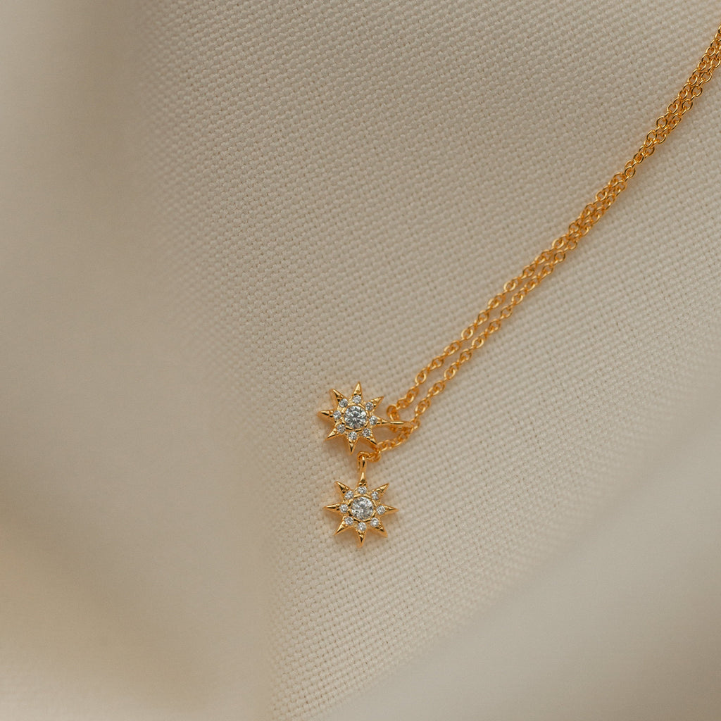 Lux Threader Earrings—gold earrings with star-shaped charms and clear gemstones, displayed on cream fabric. A stylish match for your favorite celestial jewelry pieces.