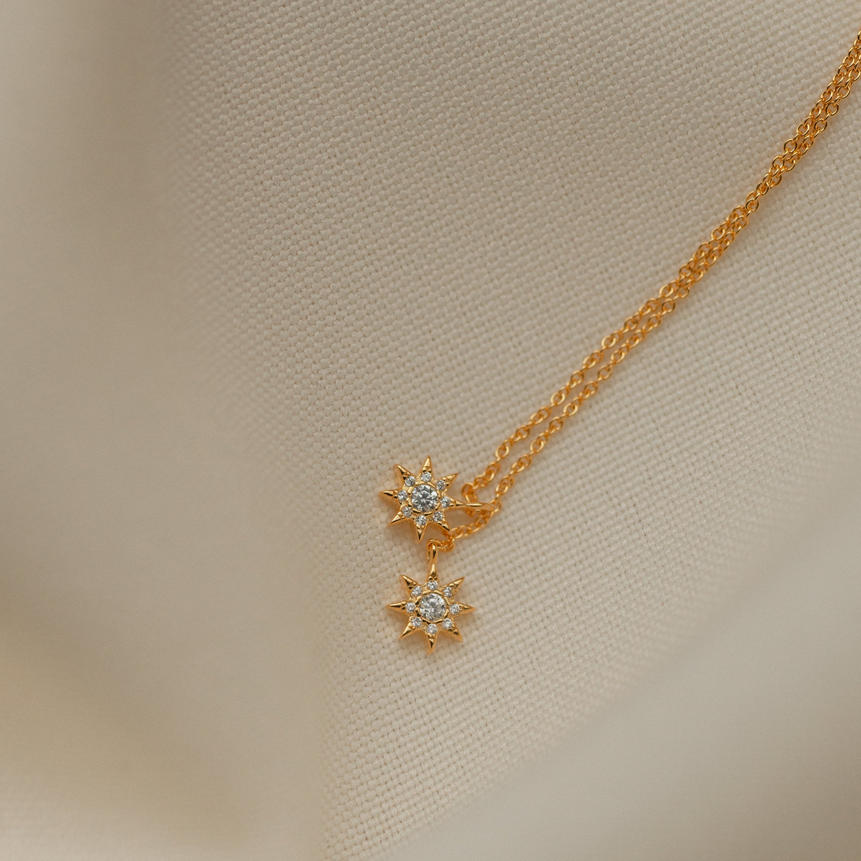 Lux Threader Earrings—gold earrings with star-shaped charms and clear gemstones, displayed on cream fabric. A stylish match for your favorite celestial jewelry pieces.