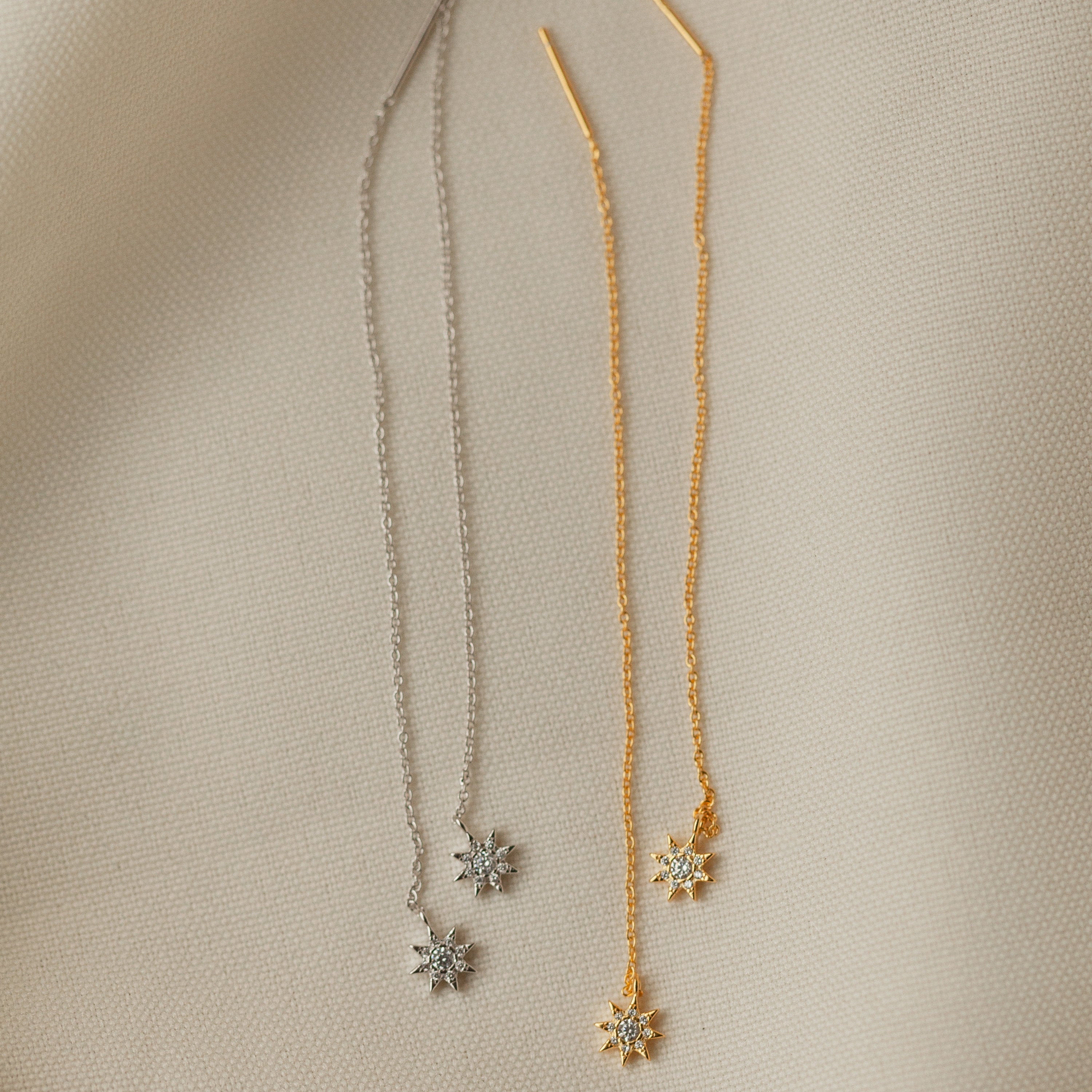 Two pairs of Lux Threader Earrings, one silver and one gold, each featuring a delicate star charm, rest on beige fabric. These chic earrings add a subtle celestial accent to any outfit.