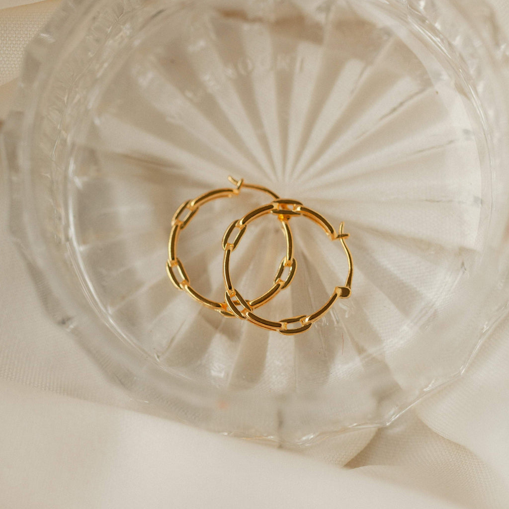 The Adele Link Hoops, featuring modern textured gold links, are displayed in a clear faceted glass dish atop a cream fabric background.