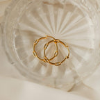 The Adele Link Hoops, featuring modern textured gold links, are displayed in a clear faceted glass dish atop a cream fabric background.