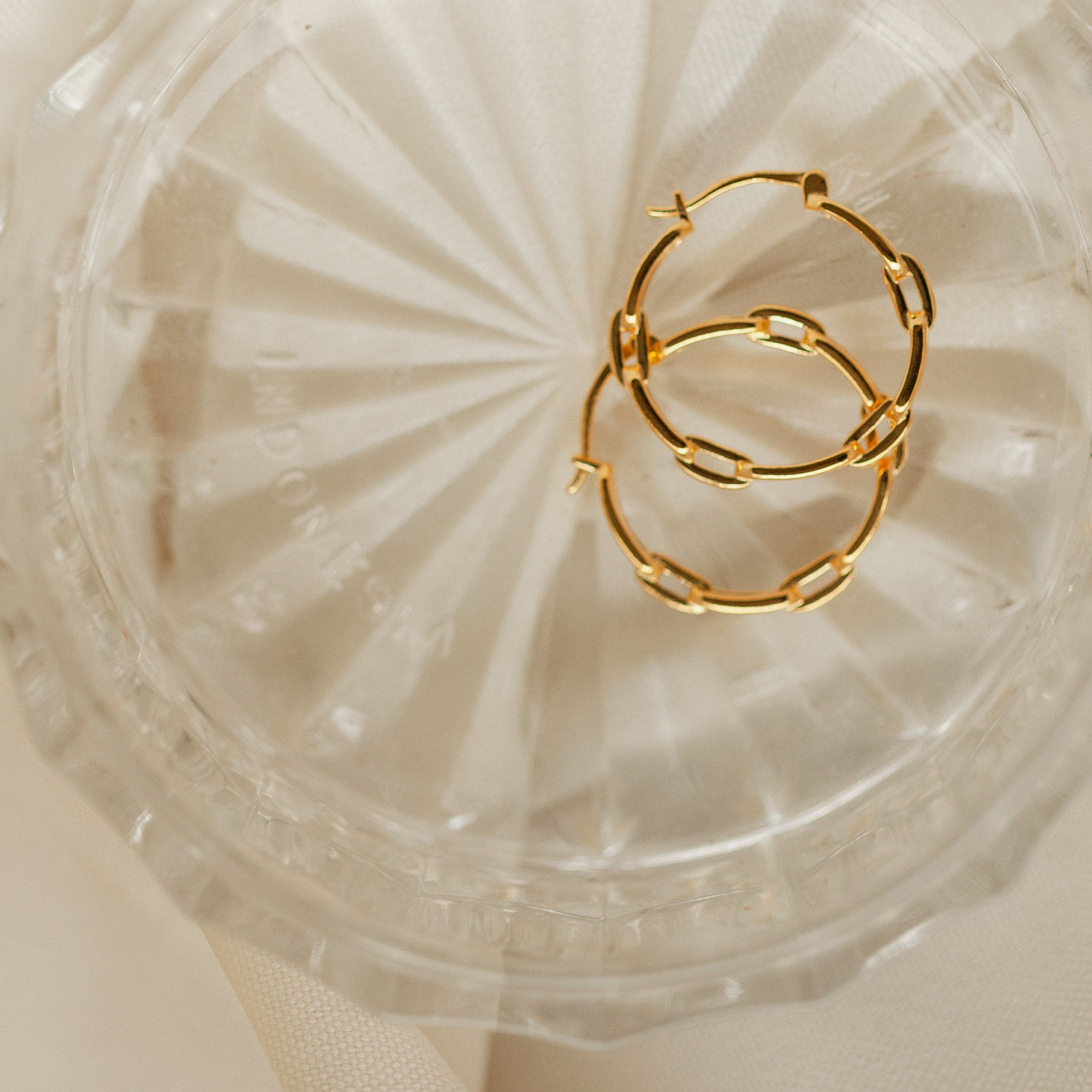 The Adele Link Hoops, featuring modern textured gold links, are displayed in a clear, textured glass dish against a light background.
