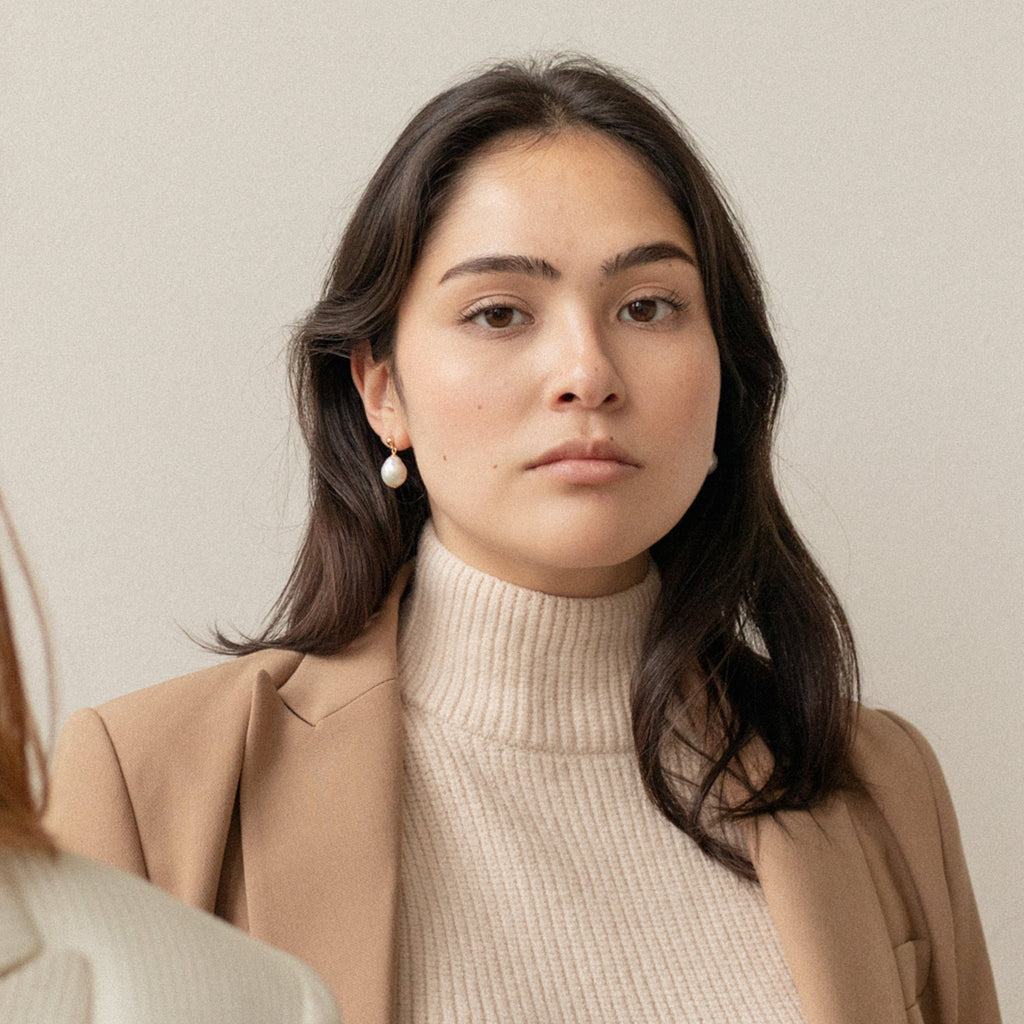A woman with long dark hair in a beige turtleneck and blazer stands against a light background, wearing elegant Baroque Pearl Earrings as she looks forward.