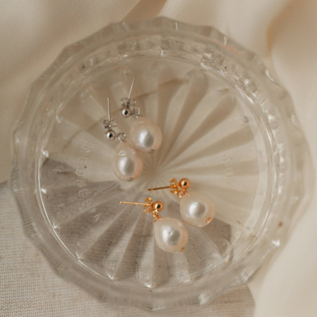 Two pairs of Baroque Pearl Earrings—one classic and one teardrop style, featuring silver and gold posts—are elegantly displayed on a clear glass dish.