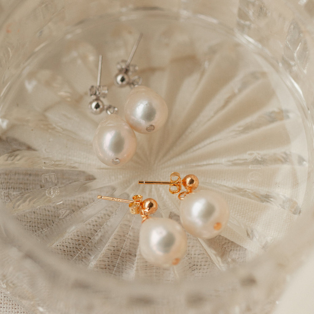 Two pairs of Baroque Pearl Earrings—one with gold backs and one with silver backs—are elegantly displayed in a clear glass dish.