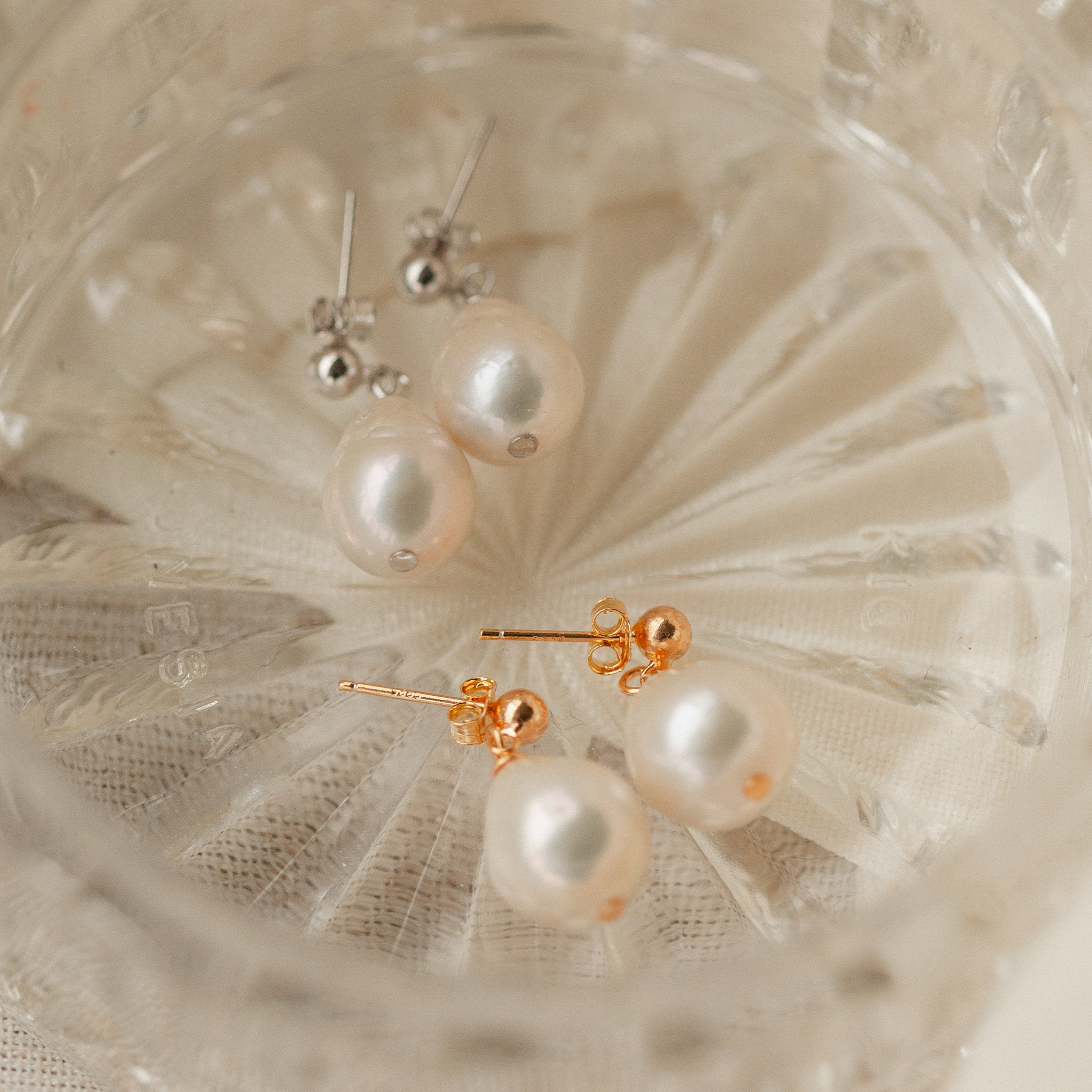 Two pairs of Baroque Pearl Earrings—one with gold backs and one with silver backs—are elegantly displayed in a clear glass dish.