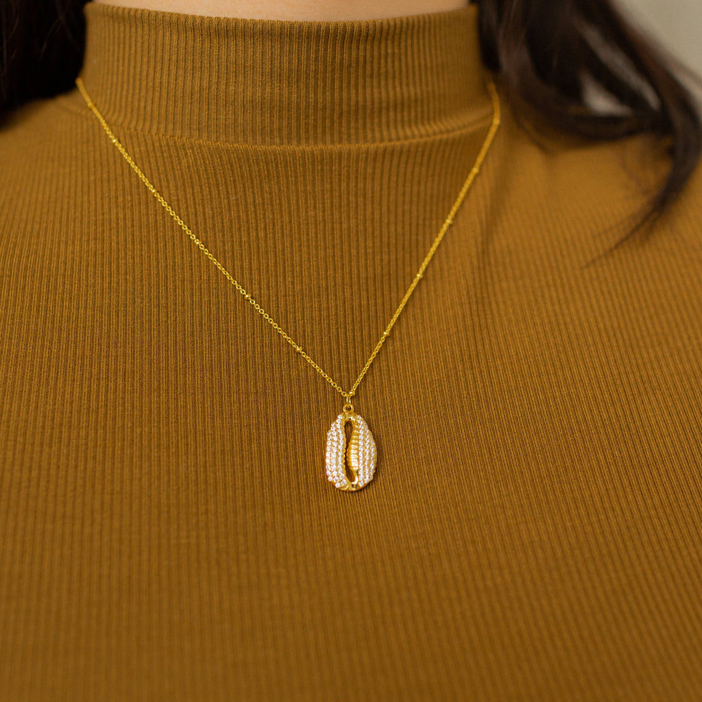 The Pave Shell Necklace on a gold chain, styled over a ribbed brown turtleneck, brings a touch of coastal cottage charm to your outfit.