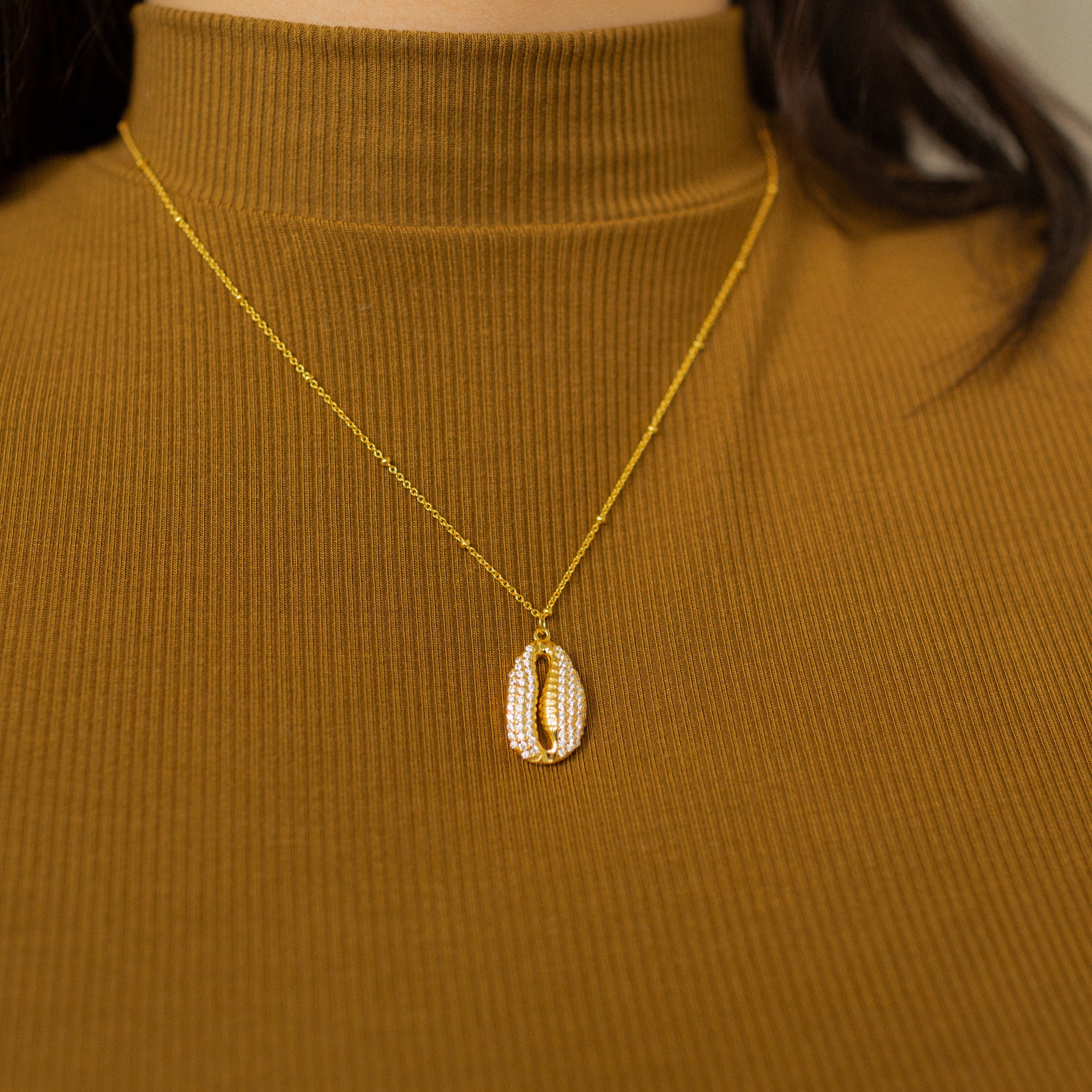 The Pave Shell Necklace on a gold chain, styled over a ribbed brown turtleneck, brings a touch of coastal cottage charm to your outfit.