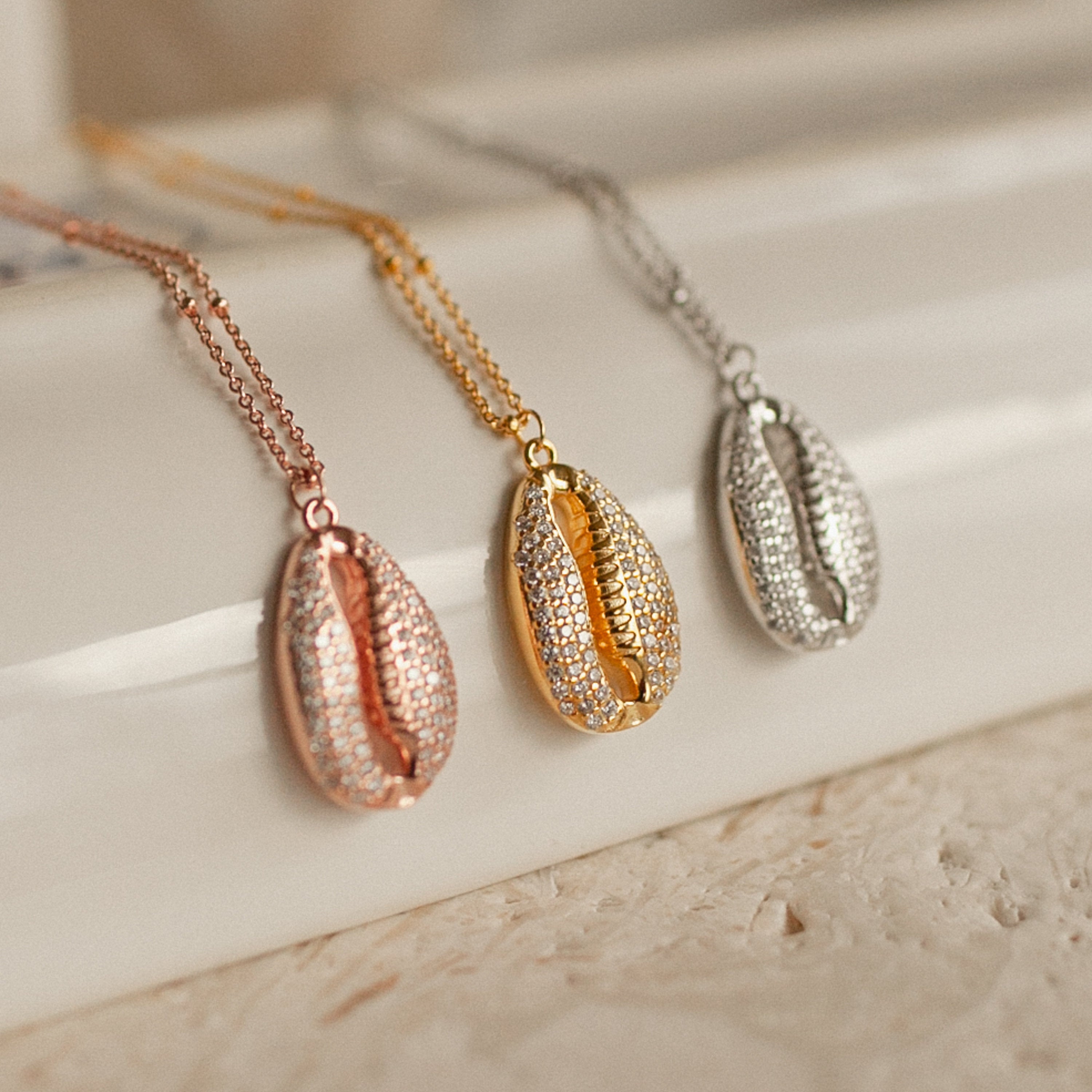 The Pave Shell Necklace, available in rose gold, gold, and silver, adds coastal cottage charm to any outfit and looks stunning when displayed on a light, textured surface.