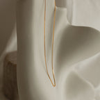 A single gold box chain necklace hangs vertically across the contours of an abstract cream sculpture,