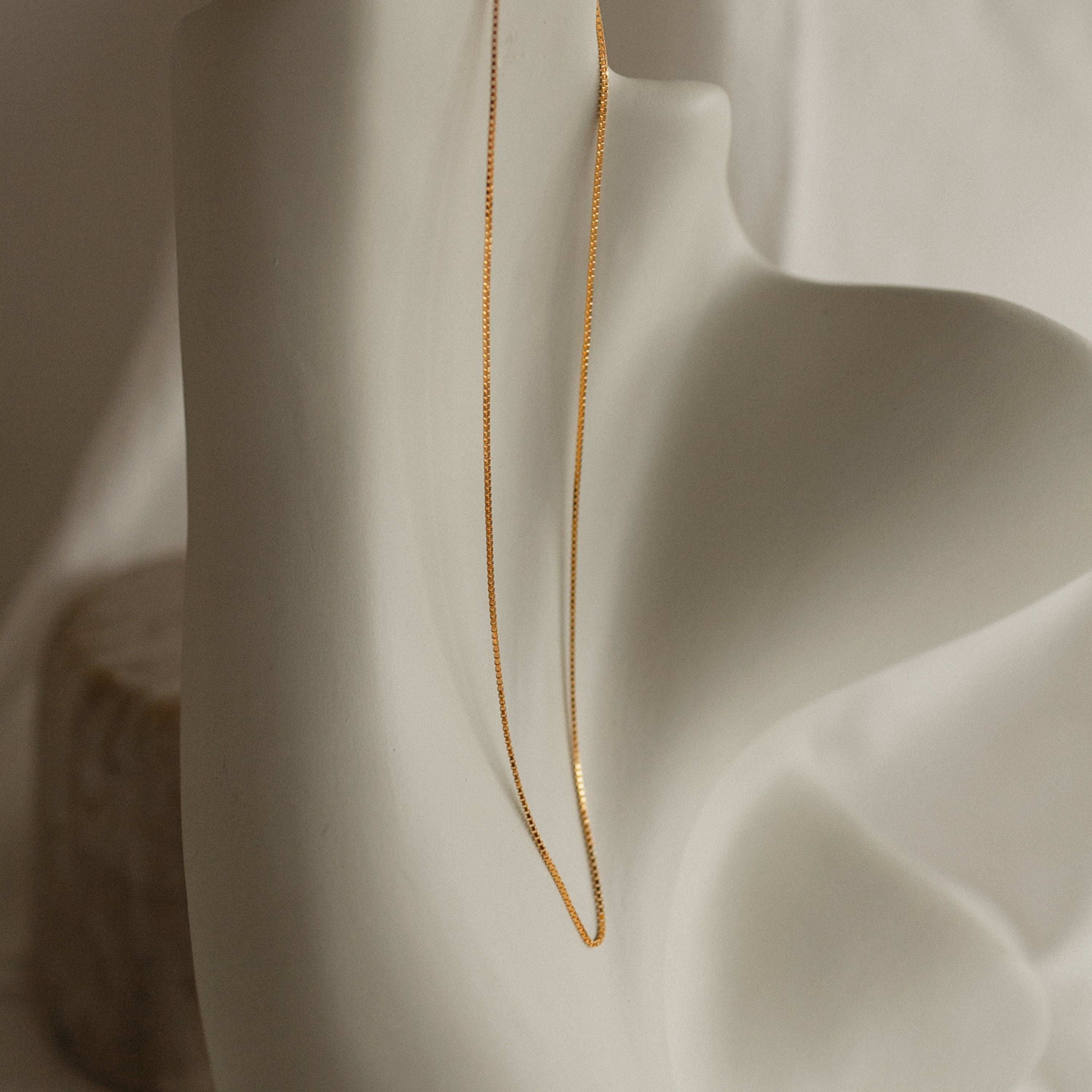 A single gold box chain necklace hangs vertically across the contours of an abstract cream sculpture,