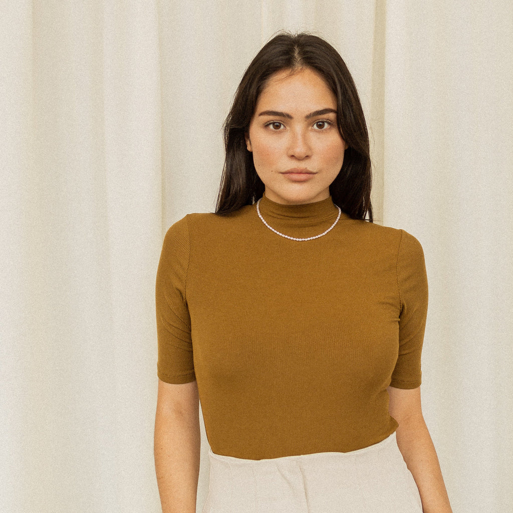 A woman with long dark hair, dressed in a brown top, wears the Pearl Choker Necklace while standing before cream-colored curtains.