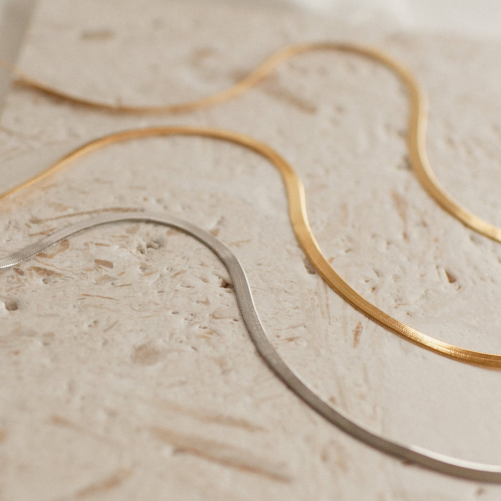 Two yellow gold herringbone necklaces and one white gold herringbone necklace laid flat on a beige stone surface.