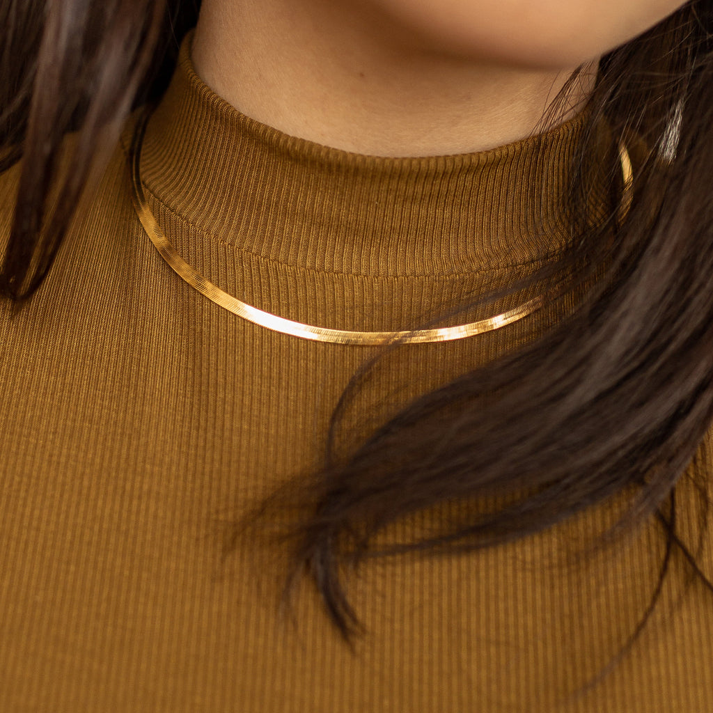 A close-up of a gold herringbone necklace worn over a brown ribbed high-neck top, with dark hair partially covering the view.