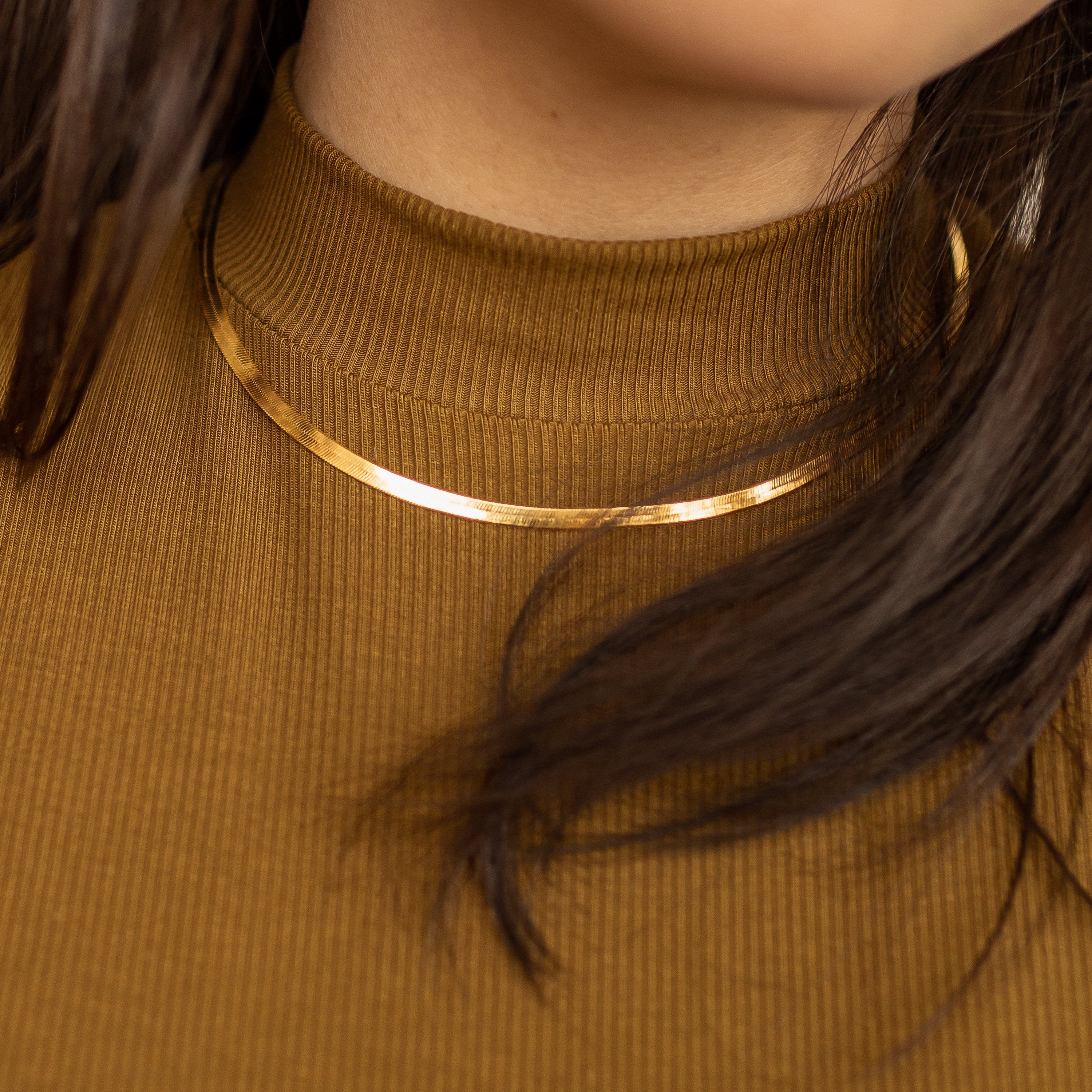 A close-up of a gold herringbone necklace worn over a brown ribbed high-neck top, with dark hair partially covering the view.