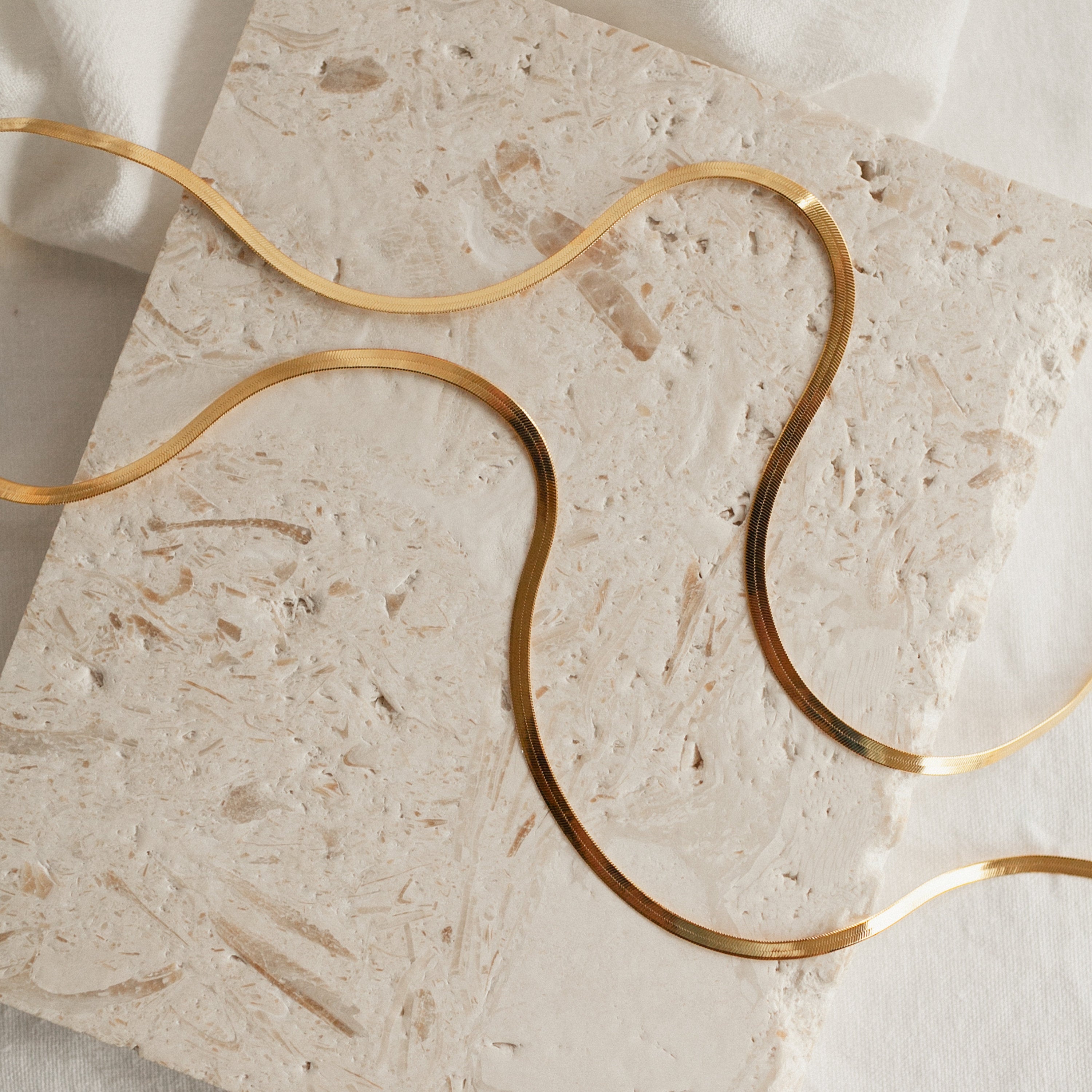 Two yellow gold herringbone necklaces arranged in wavy curves on a light, textured surface.