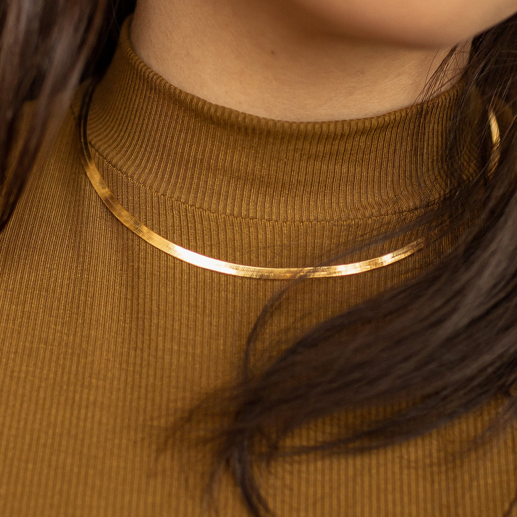 A gold herringbone necklace worn over a brown ribbed turtleneck top.