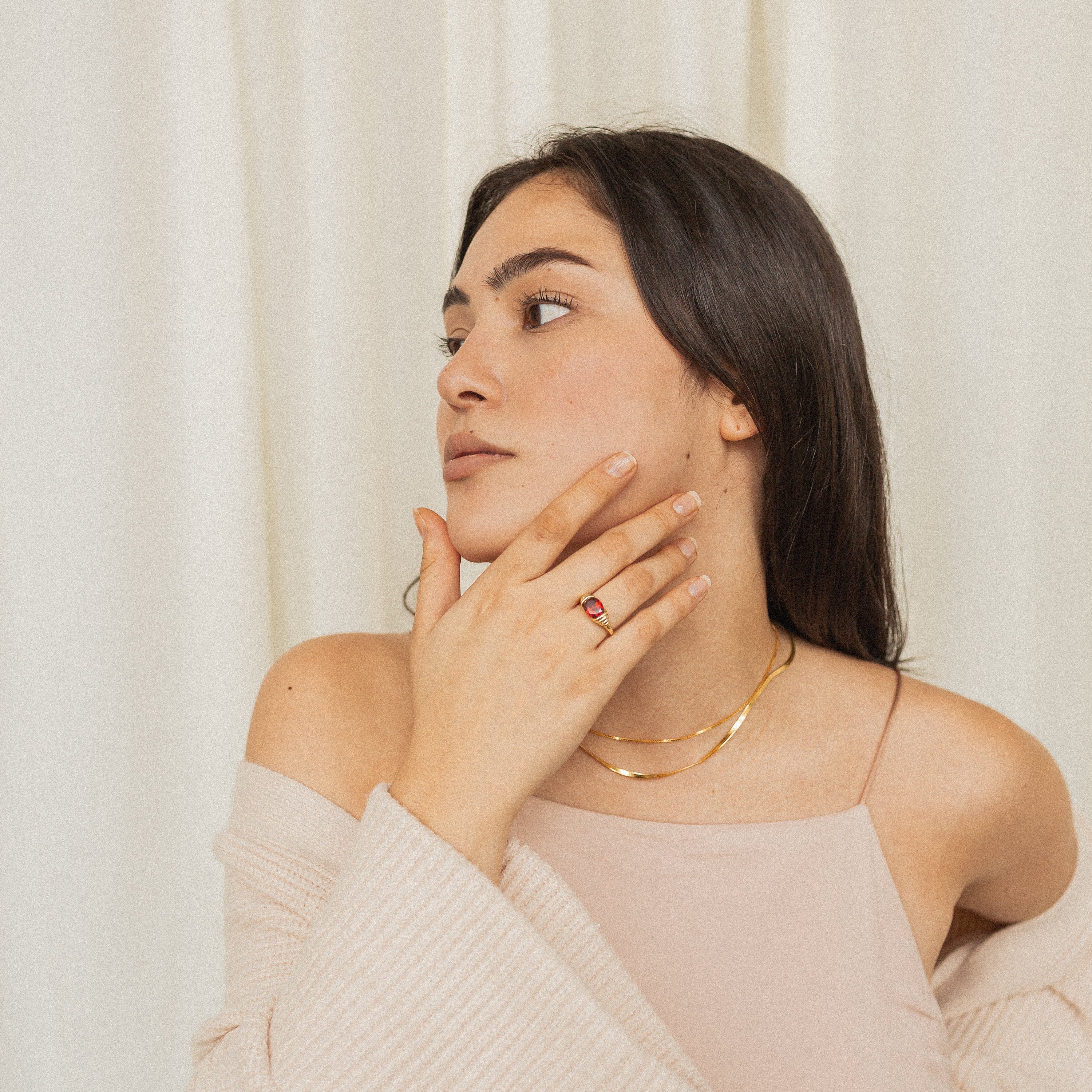 A woman with long dark hair touches her neck, wearing a gold necklace and the Garnet Signet Ring, set against a cream background.