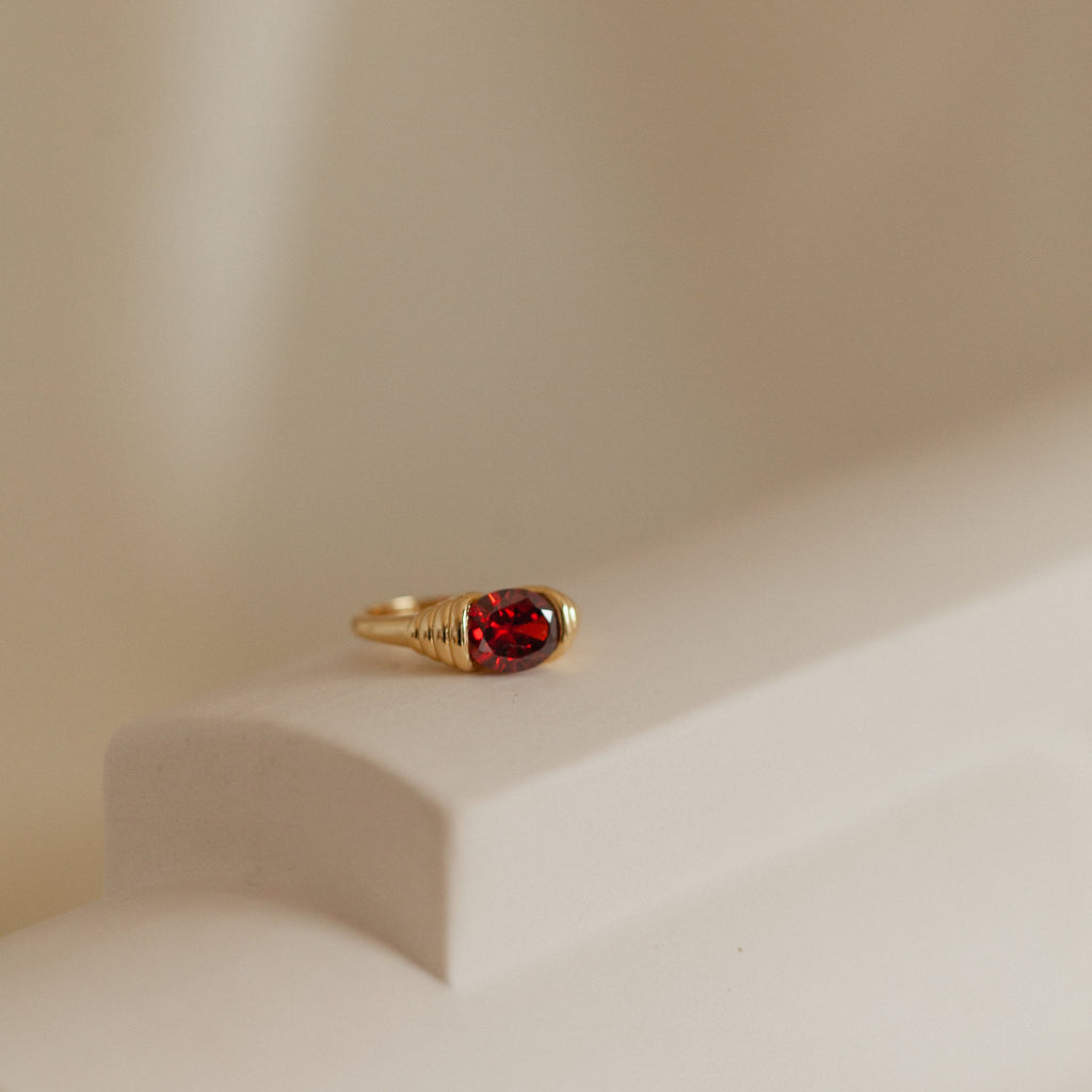 The Garnet Signet Ring, featuring a red gemstone, is showcased on a smooth beige surface with soft lighting—an elegant and timeless signature jewelry piece.