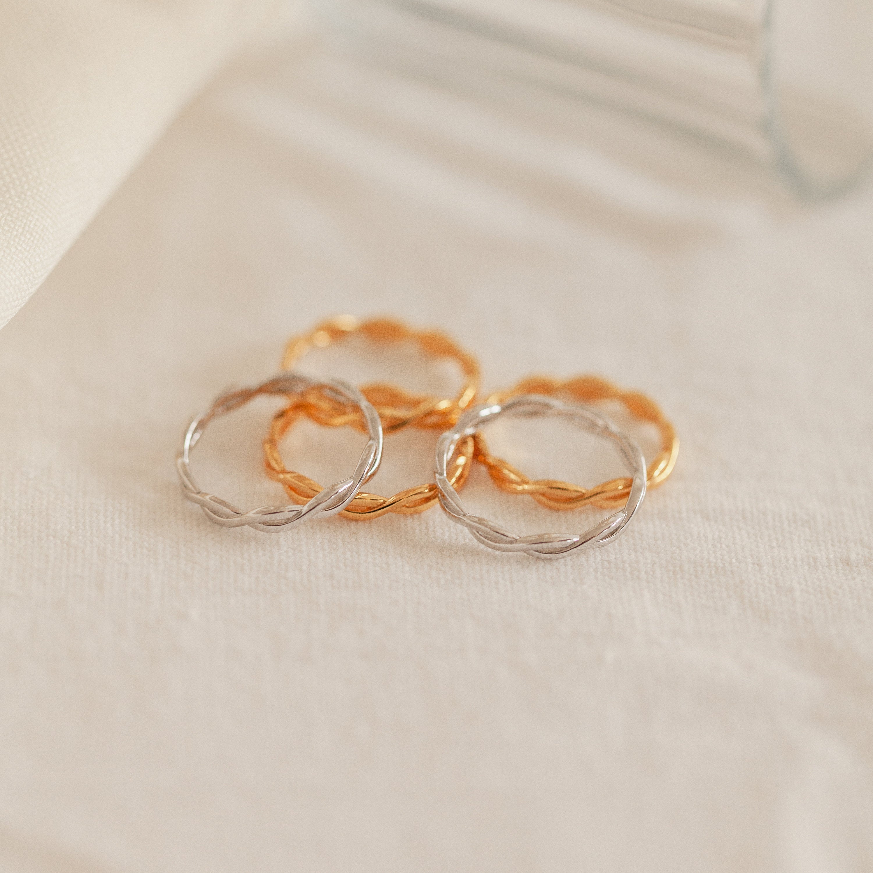 The Braided Ring and three other twisted metal rings, including a sleek silver band, are displayed on a soft, cream-colored fabric surface.