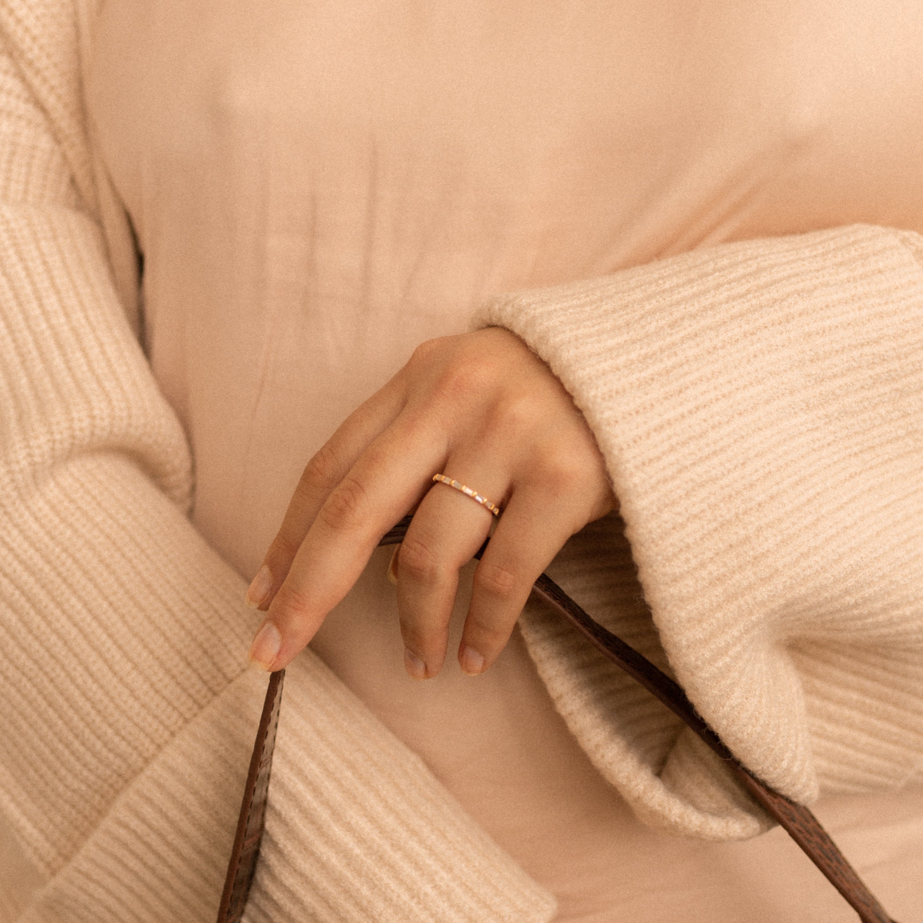 A hand wearing the Baguette Eternity Ring holds a brown strap while dressed in a cream ribbed sweater and beige top.