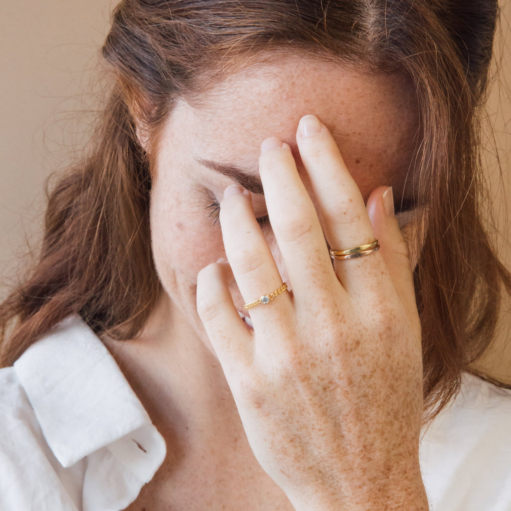 A woman with freckles covers part of her face with her hand, showcasing the edgy Birthstone Curb Ring while wearing a white shirt.