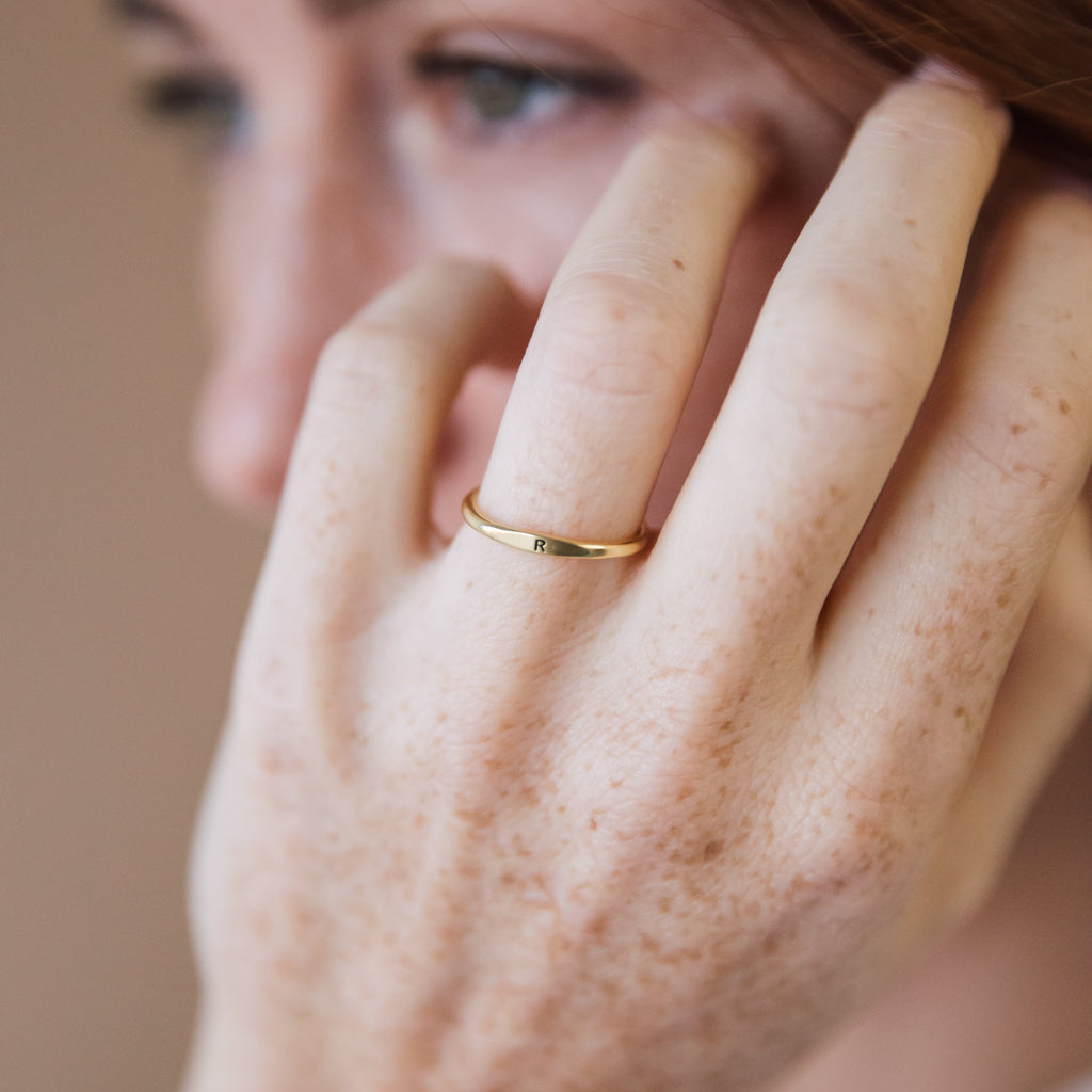 Close-up of a freckled hand wearing the Skinny Initial Ring—a gold, personalized dainty ring engraved with “R,” perfect for stacking.