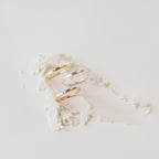 The Skinny Initial Ring, engraved and available in gold, silver, and rose gold, is displayed stacked on white textured powder against a light background.