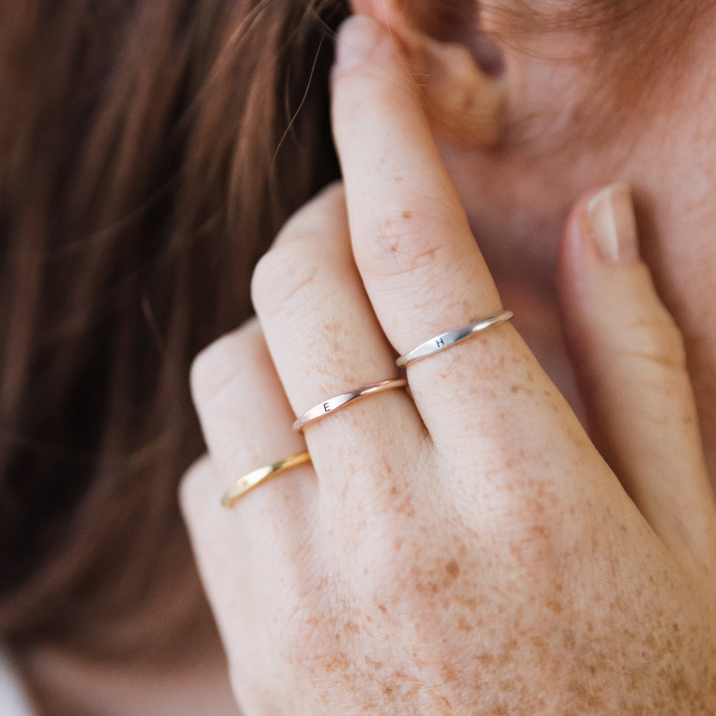 A hand with three dainty Skinny Initial Rings—gold, rose gold, and silver—stacked on freckled fingers near the ear, perfect for layering or personalizing your look.