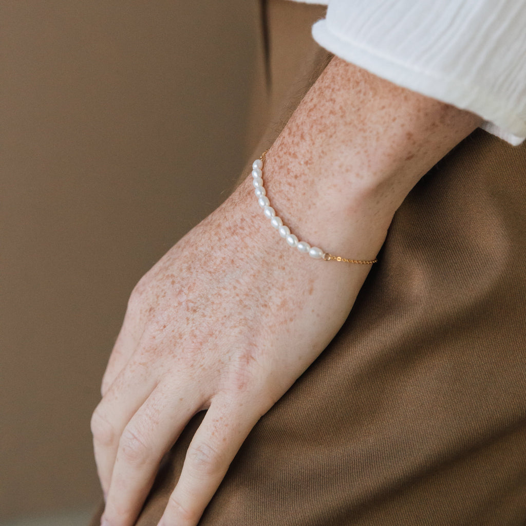 A hand adorned with the Pearl Bead Bracelet, featuring modern pearls and gold, rests on brown pants, showcasing the bracelet’s delicate elegance.