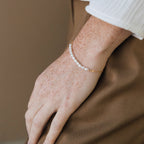 A hand adorned with the Pearl Bead Bracelet, featuring modern pearls and gold, rests on brown pants, showcasing the bracelet’s delicate elegance.