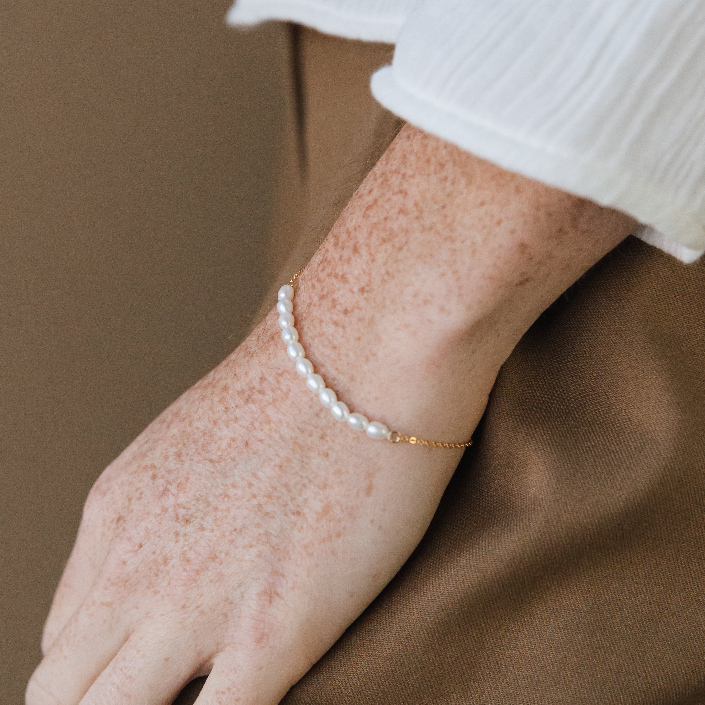 A hand adorned with the Pearl Bead Bracelet, featuring modern pearls and a gold chain, rests gracefully on a brown fabric surface.