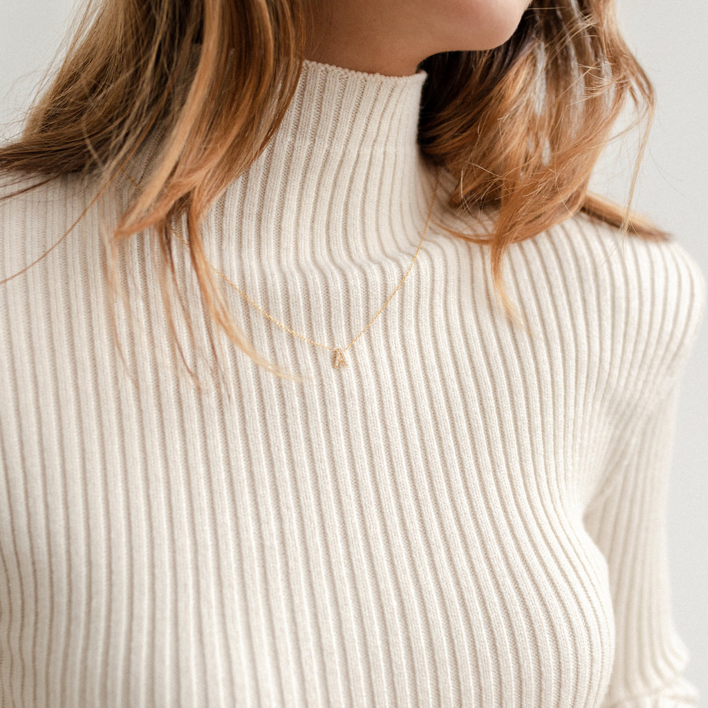 A woman wears a cream ribbed turtleneck sweater and the Pave Initial Necklace in Rose Gold, adding an elegant, personalized touch to her look.