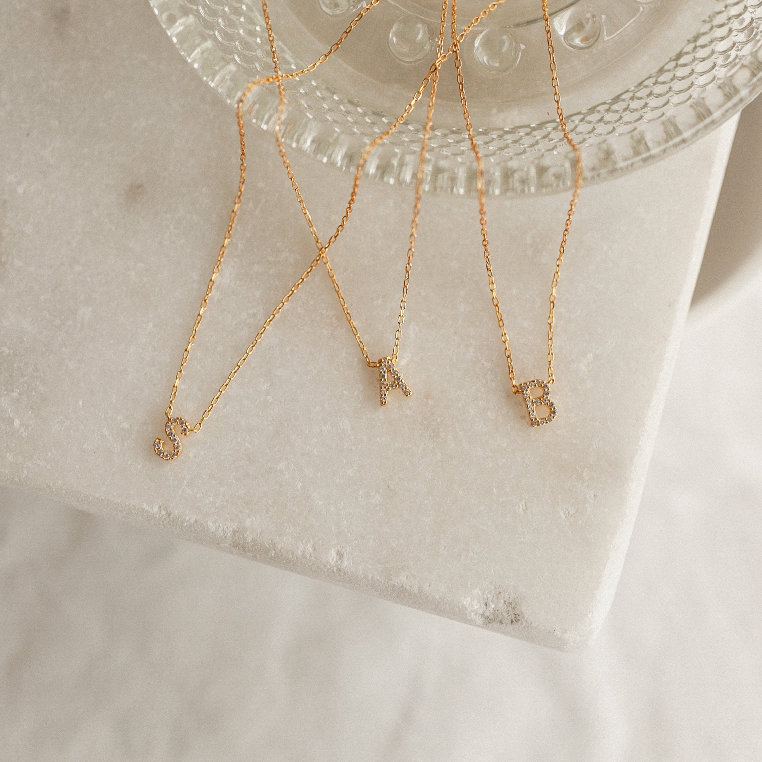 Three Pave Initial Necklaces featuring gold letter pendants J, A, and B are displayed on a marble surface by a glass dish—an elegant showcase of personalized jewelry.