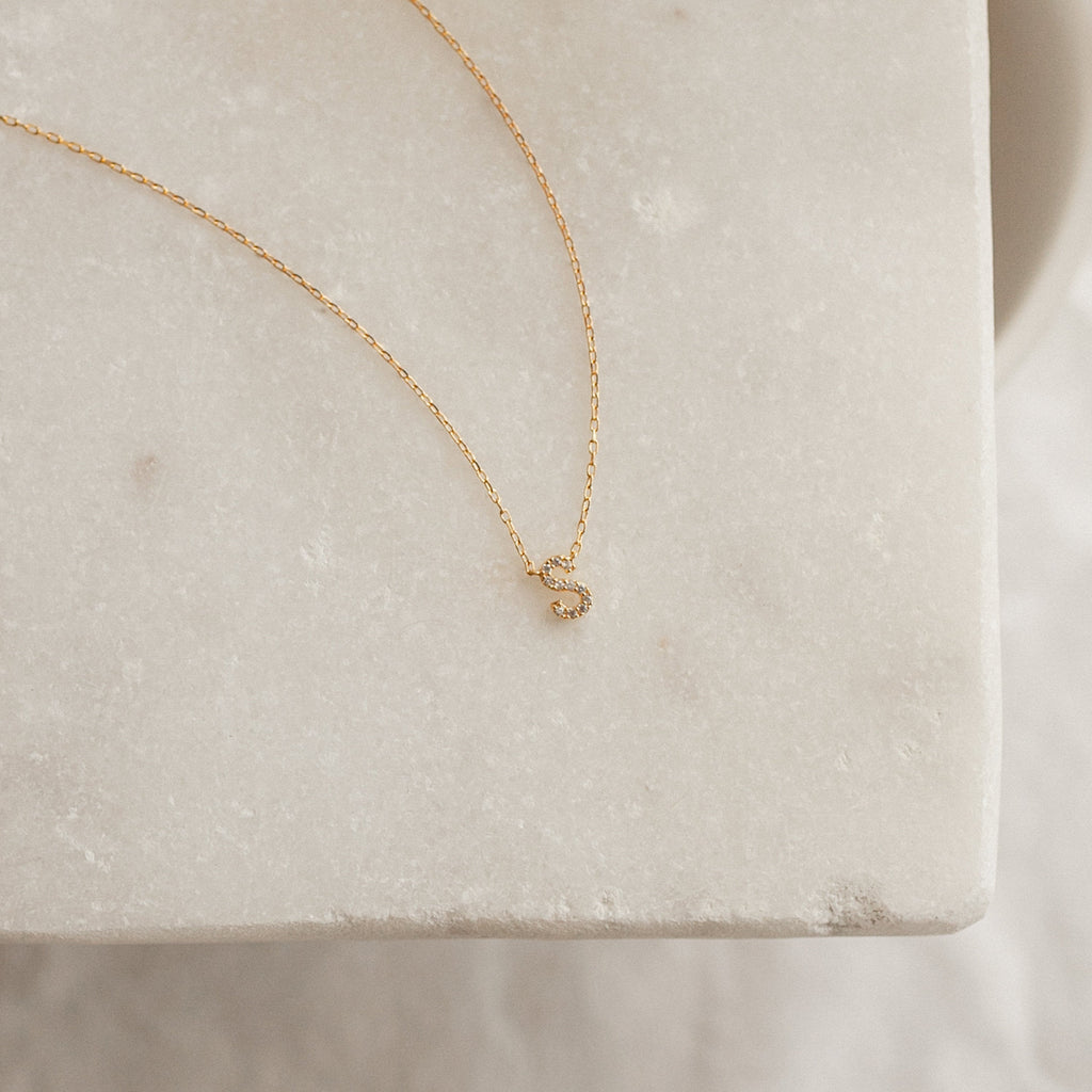 The Pave Initial Necklace features a small gold letter "S" pendant on a chic chain, displayed on white marble—a stylish personalized piece perfect for everyday wear.