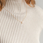The Pave Initial Necklace, featuring a sparkling gold letter A pendant, shines on a ribbed cream turtleneck sweater, adding personalized charm to the look.