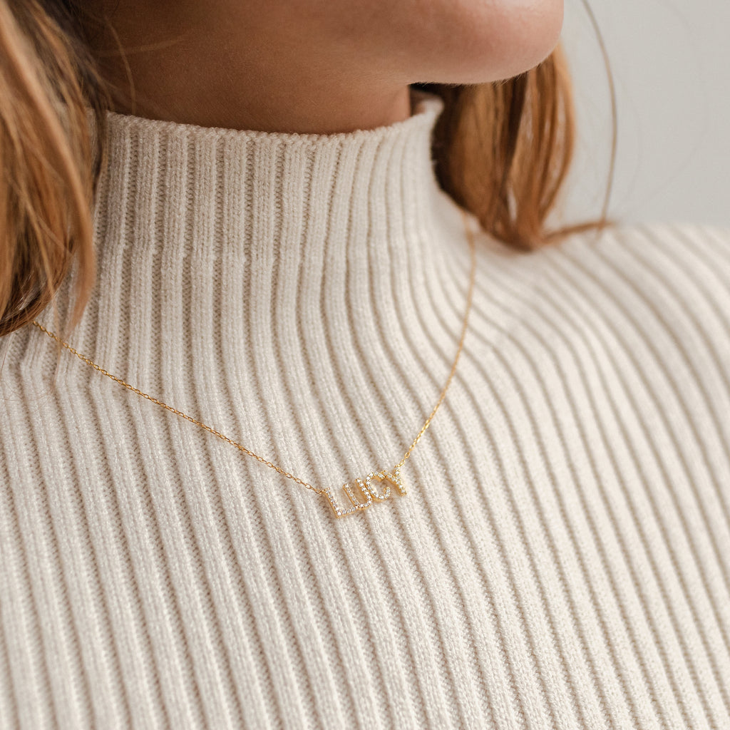 Close-up of a woman wearing the Pave Name Necklace with personalized block letters, styled over a cream ribbed turtleneck sweater.