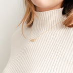 A woman in a cream ribbed turtleneck wears a rose gold Pave Name Necklace featuring block letters.