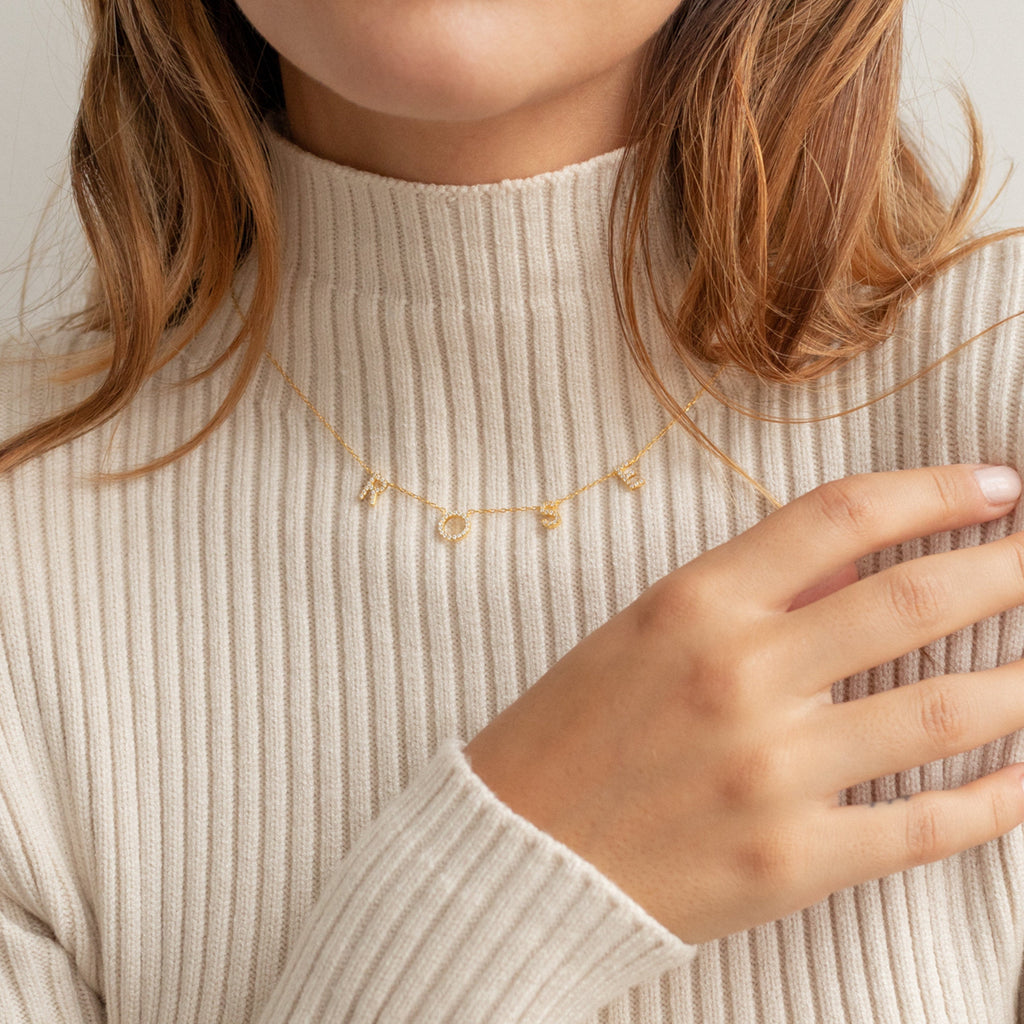 A woman in a beige ribbed turtleneck wears the Pave Letter Necklace, a personalized gold jewelry piece with LOVE charms, as she touches her collar.