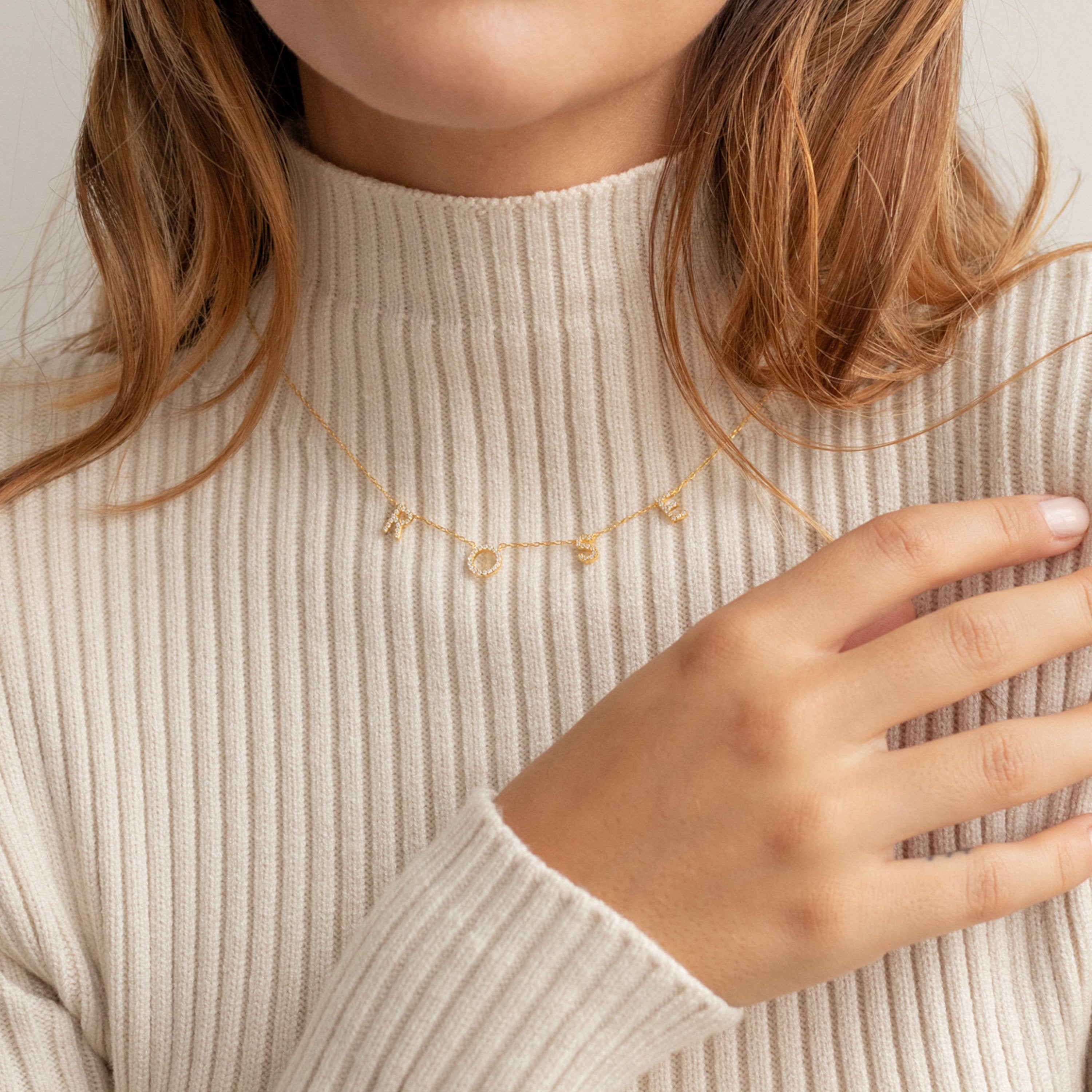 A woman in a beige ribbed turtleneck wears the Pave Letter Necklace, a personalized gold jewelry piece with LOVE charms, as she touches her collar.