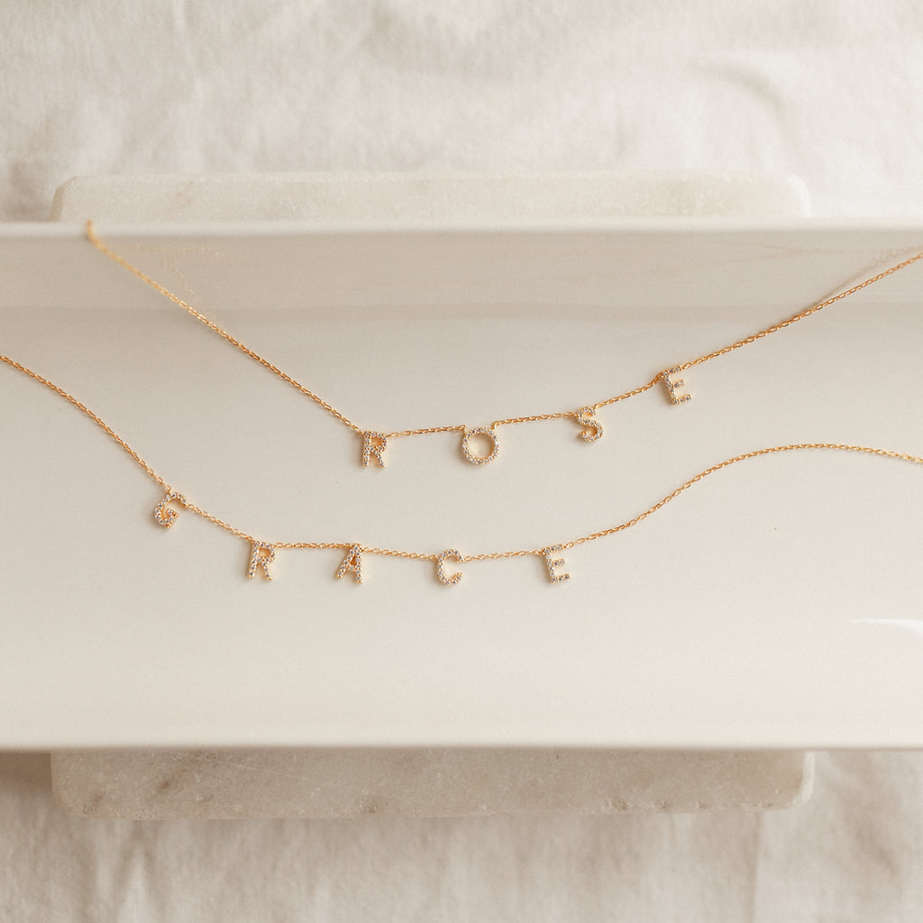 Two Pave Letter Necklaces in Rose Gold, featuring charms that spell ROSE and GRACE, are arranged on a white tray—an elegant way to personalize your style.