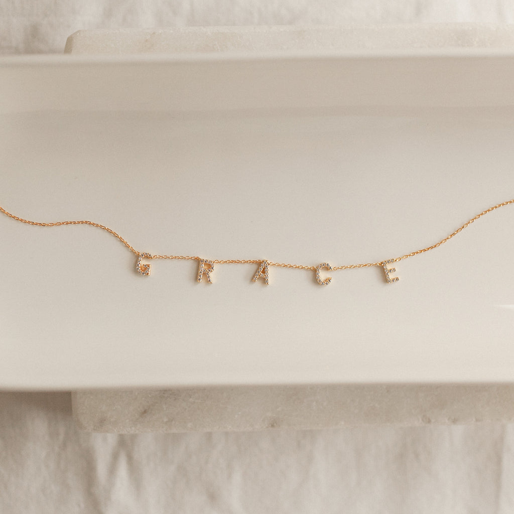 The Pave Letter Necklace features dangling letter charms spelling GRACE, shown on a white ceramic tray—a meaningful necklace and an ideal choice for personalized jewelry.