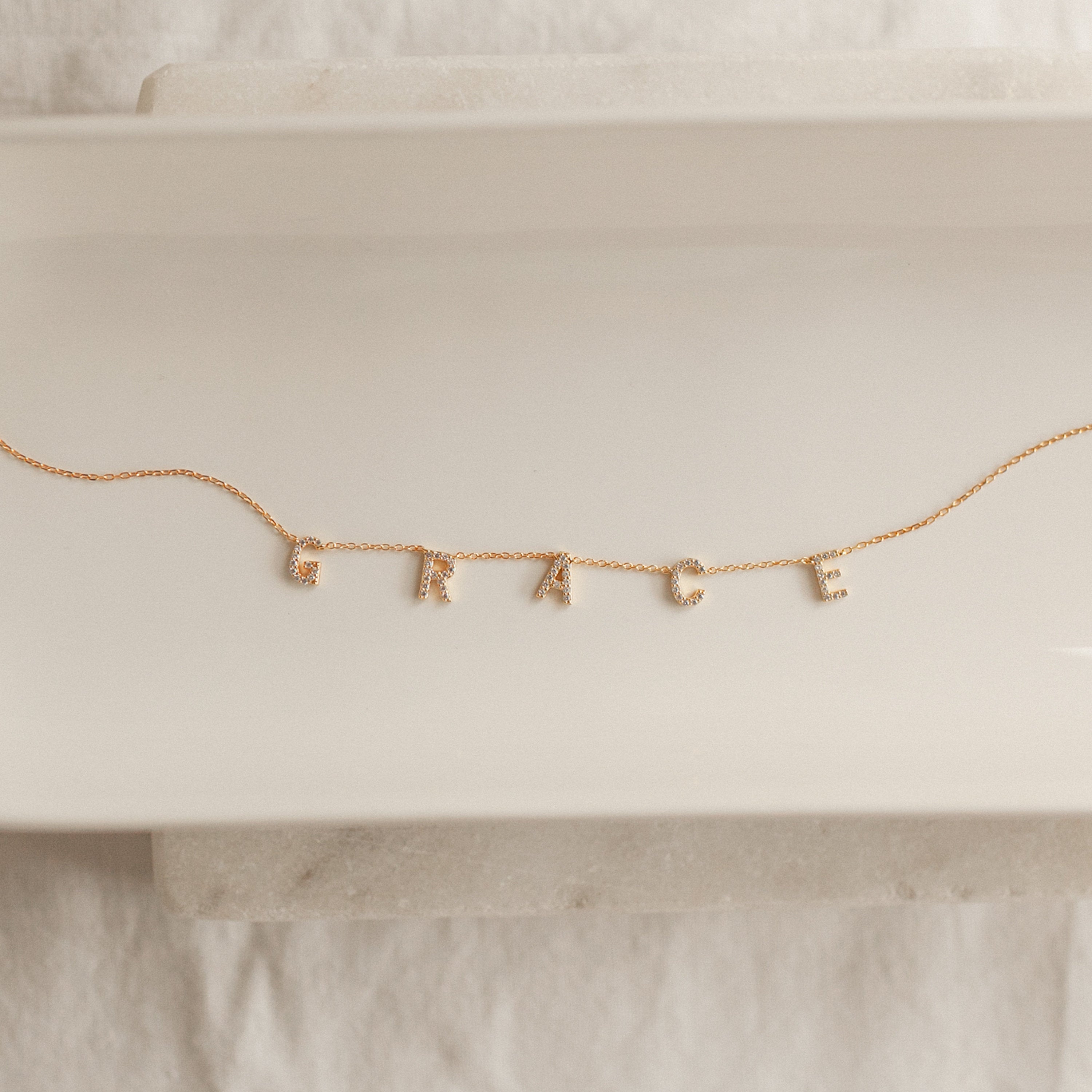 The Pave Letter Necklace features dangling letter charms spelling GRACE, shown on a white ceramic tray—a meaningful necklace and an ideal choice for personalized jewelry.