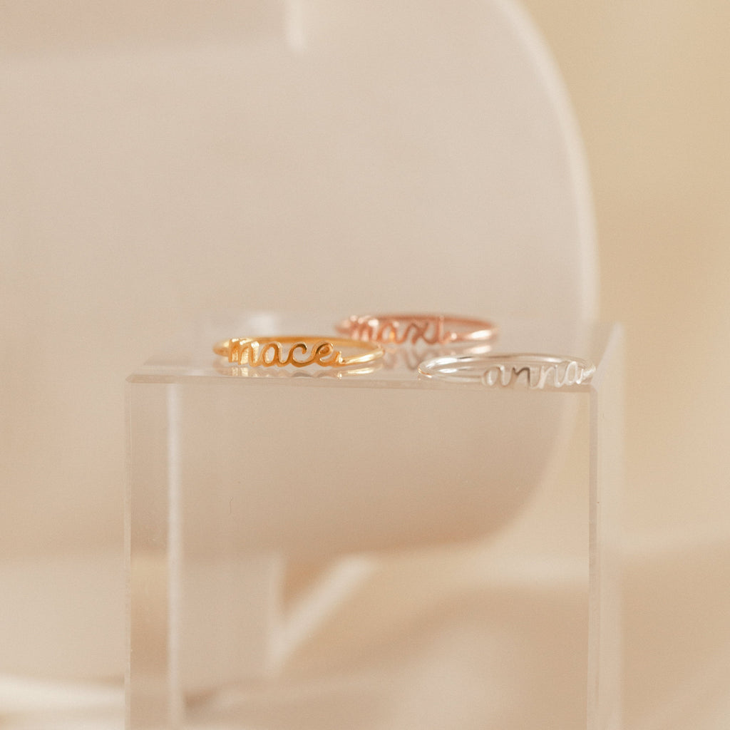 Three delicate Petite Tiny Name Rings in gold, rose gold, and silver—each a personalized script name ring—beautifully displayed on a clear acrylic stand.