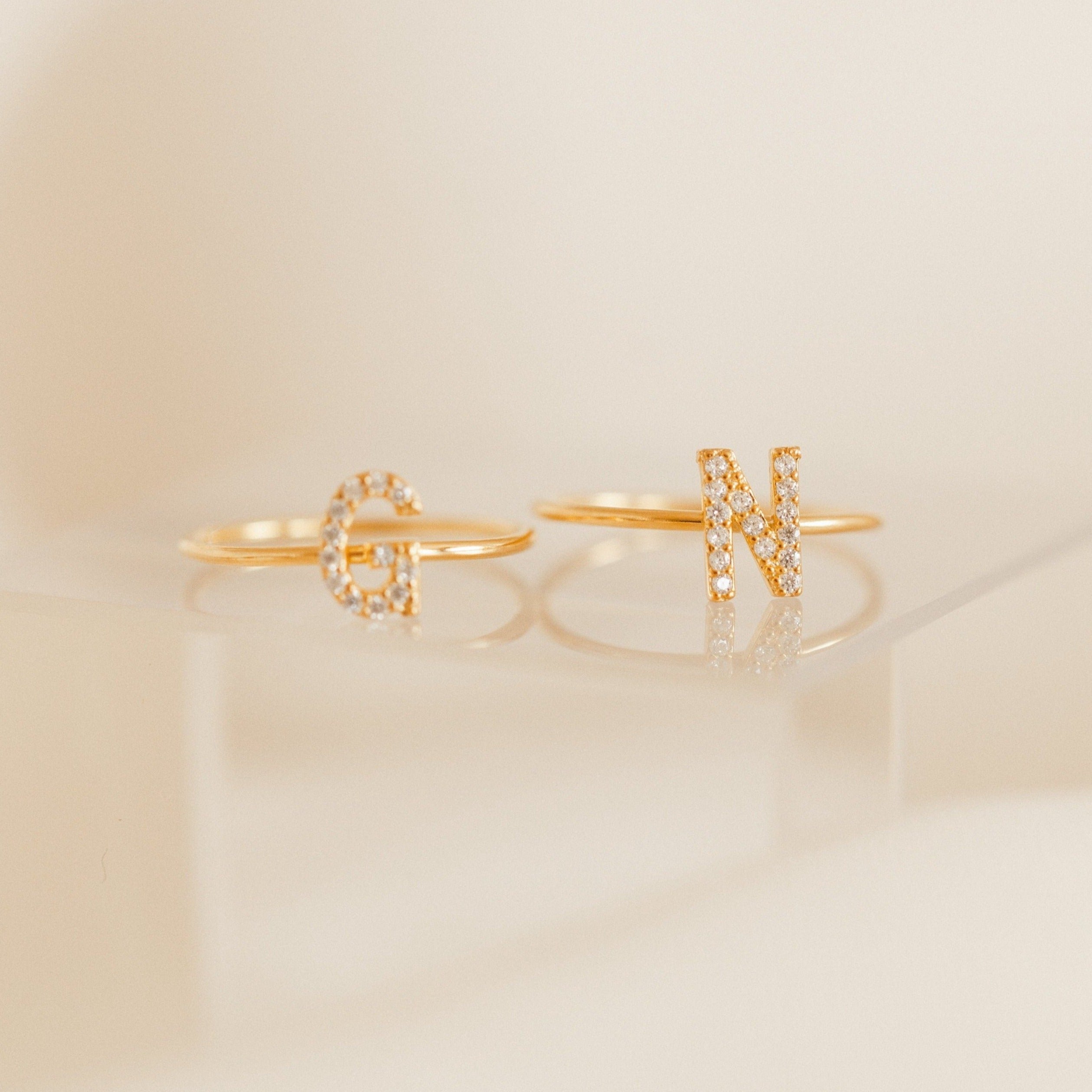 Two Pave Initial Rings, each with a diamond-encrusted "G" and "N," are displayed on a clear surface, highlighting the elegant design of this personalized jewelry.
